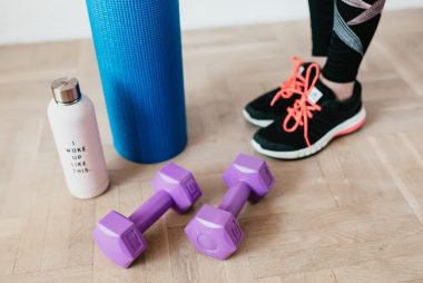 Unrecognizable sportswoman wearing sneakers and black leggings standing with sport mat on wooden floor near dumbbells and water bottle before exercising indoors